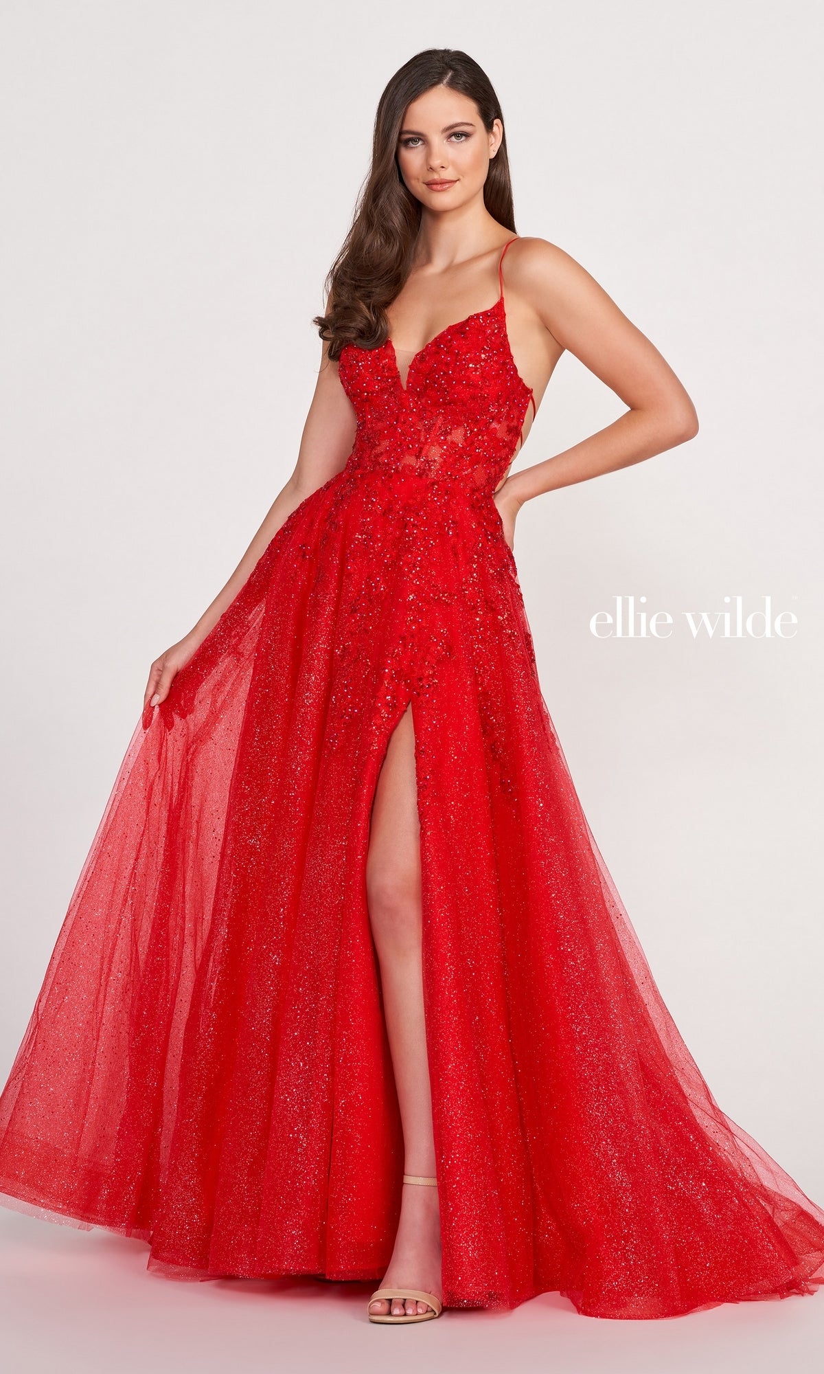 A woman models the Ellie Wilde Backless Designer Prom Dress EW34053—a sparkly red gown with a thigh-high slit and spaghetti straps—standing against a plain backdrop, the shimmering dress reflecting the light beside